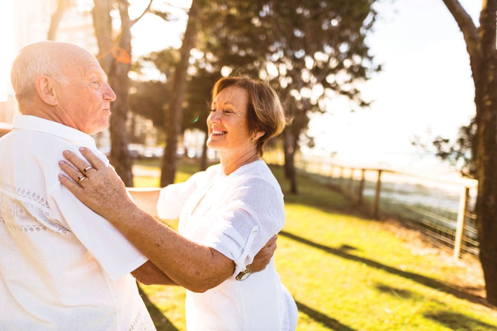 Charter Senior Living Residents couple dressed all in white smiles and dances together outdoors 